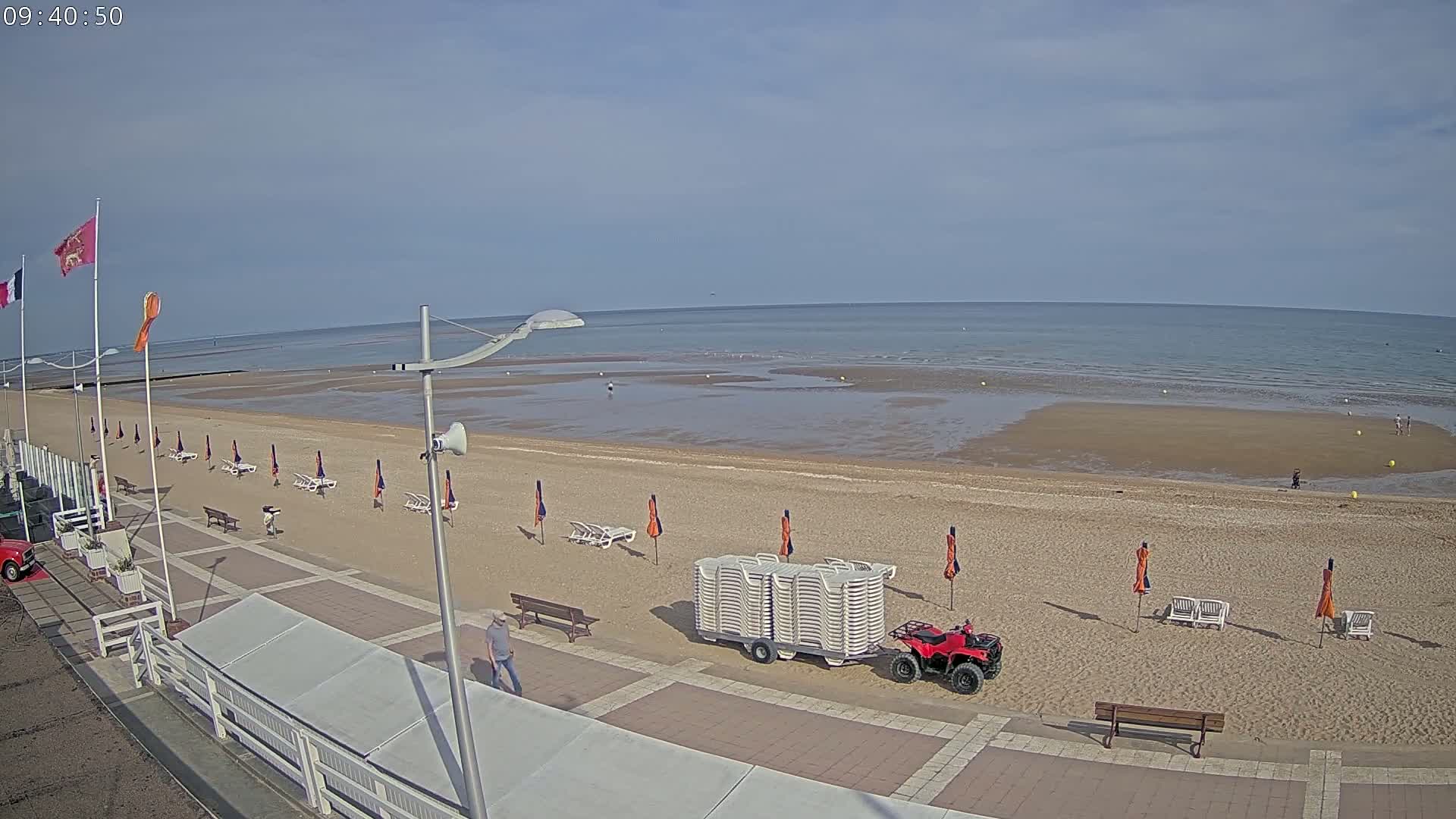 A sandy beach with a few people, beach chairs and umbrellas, a red ATV, and a stack of beach chairs on a trailer is seen on a partly sunny day next to a paved walkway.