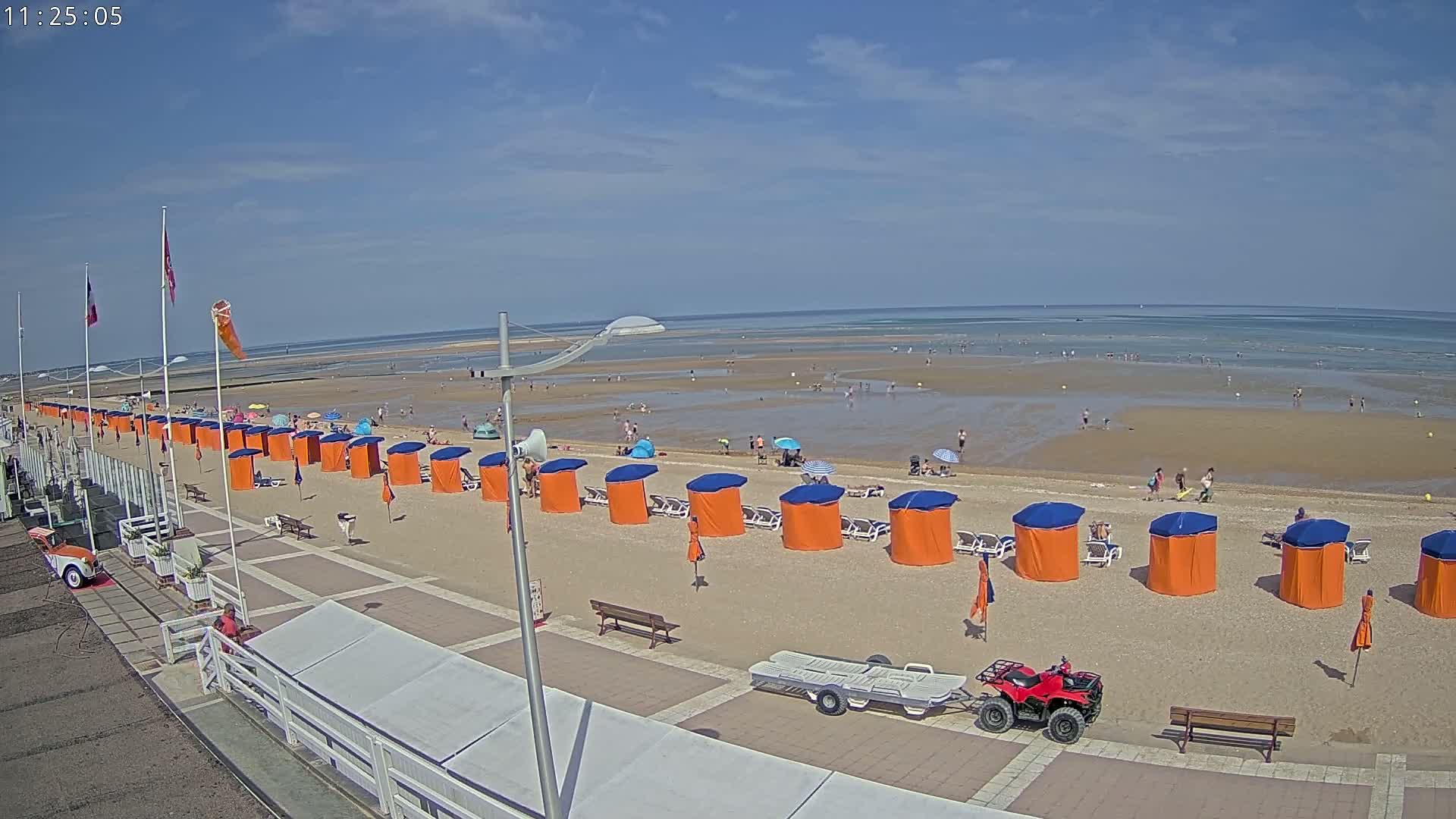 A sandy beach with a row of orange and blue beach cabanas, sparsely populated with people, stretches to a calm ocean under a clear blue sky.