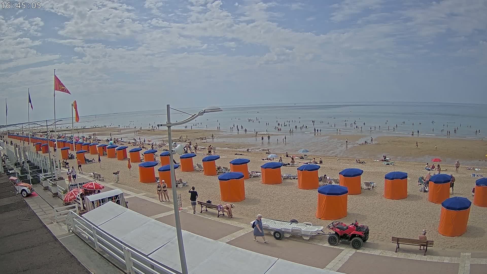 A sandy beach with many people swimming and sunbathing, lined with orange and blue beach cabanas under a partly cloudy sky.