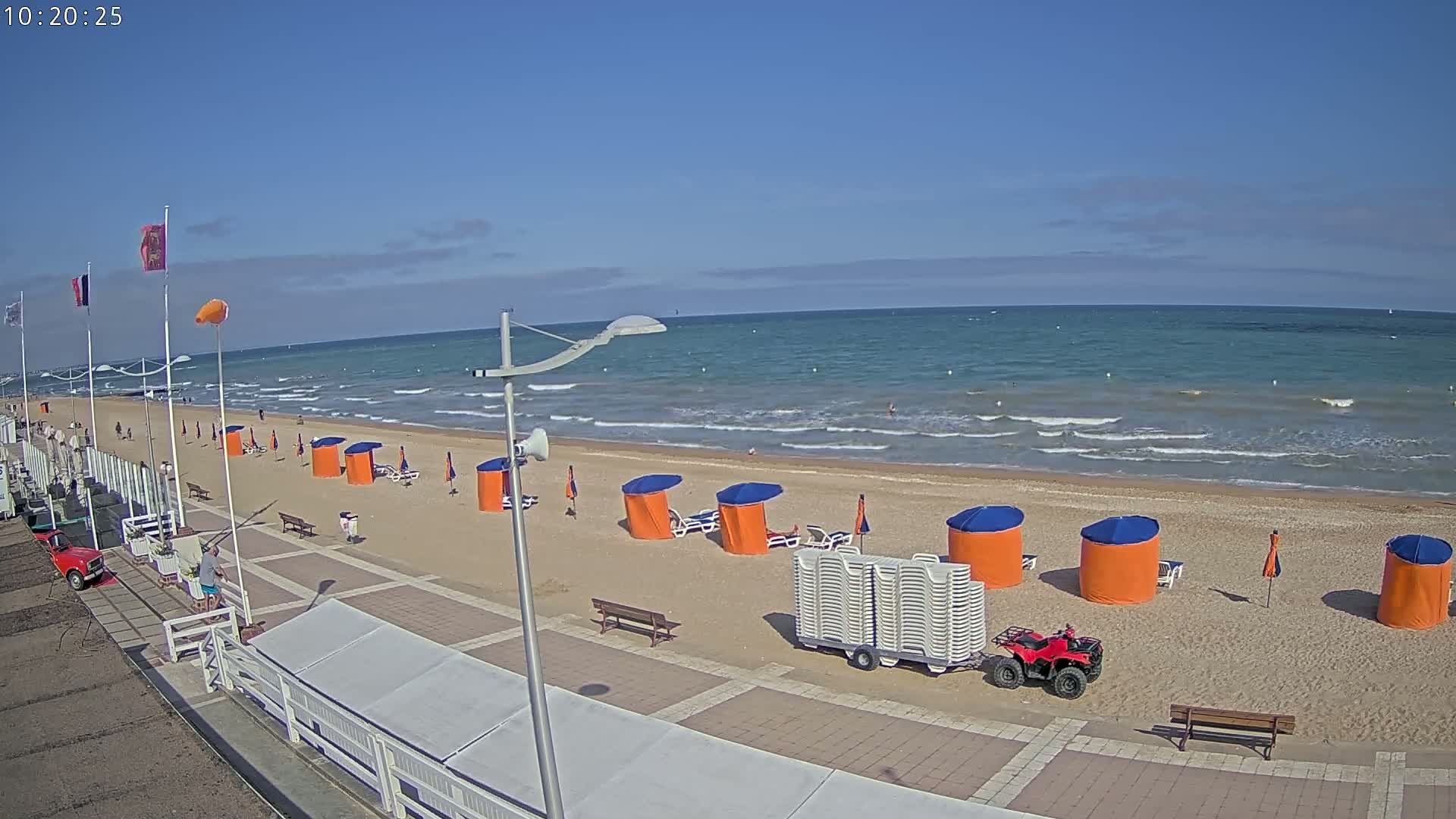 A sandy beach with small orange and blue cabanas and a red ATV is visible next to a boardwalk on a sunny day with light waves on the ocean.