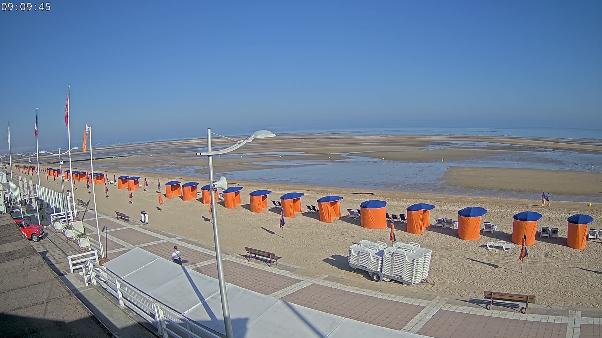 A sandy beach with rows of orange and blue beach cabanas, a boardwalk, and a calm ocean under a clear blue sky.