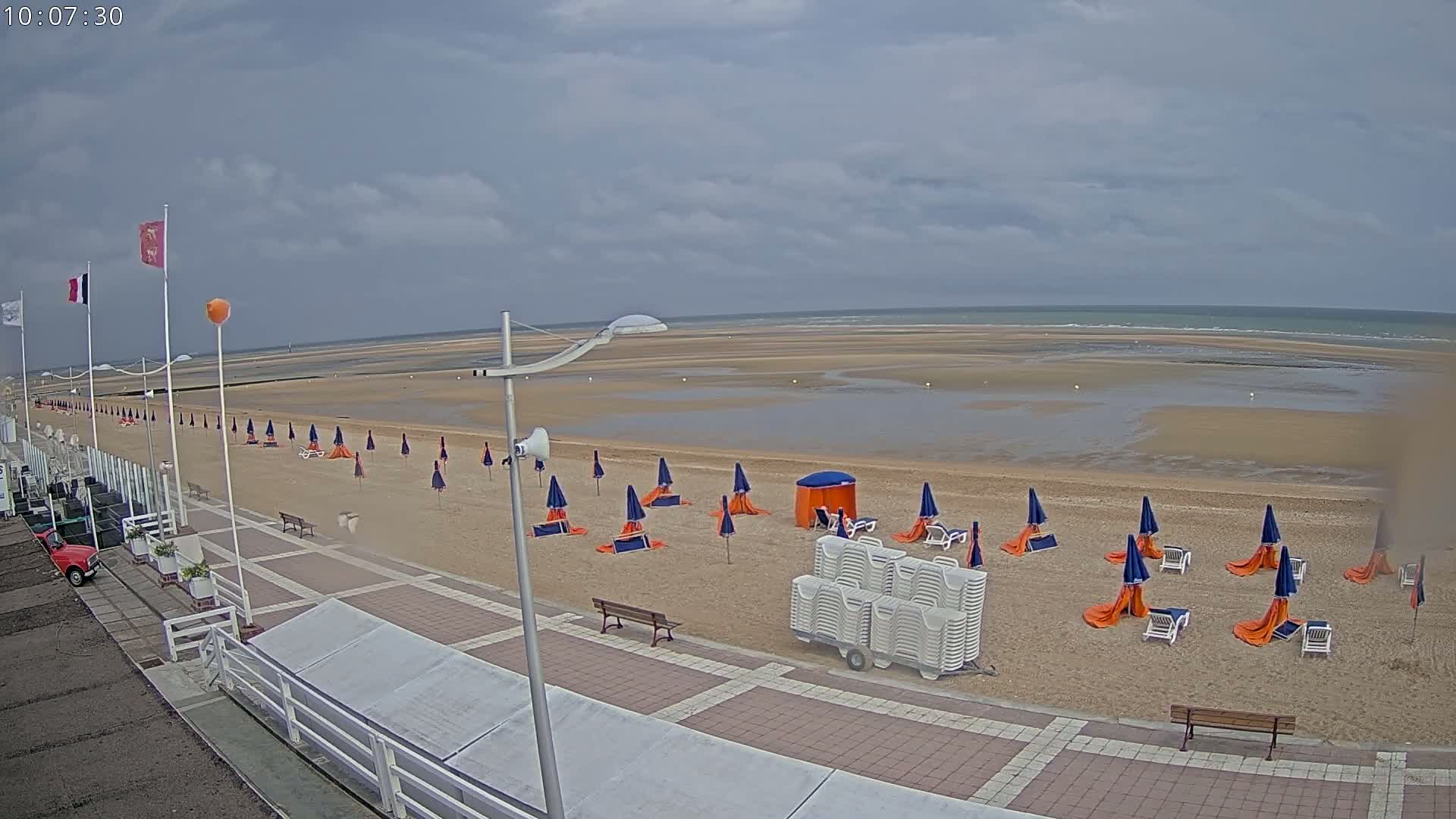 A mostly empty beach with blue and orange umbrellas and white lounge chairs is shown under an overcast sky.