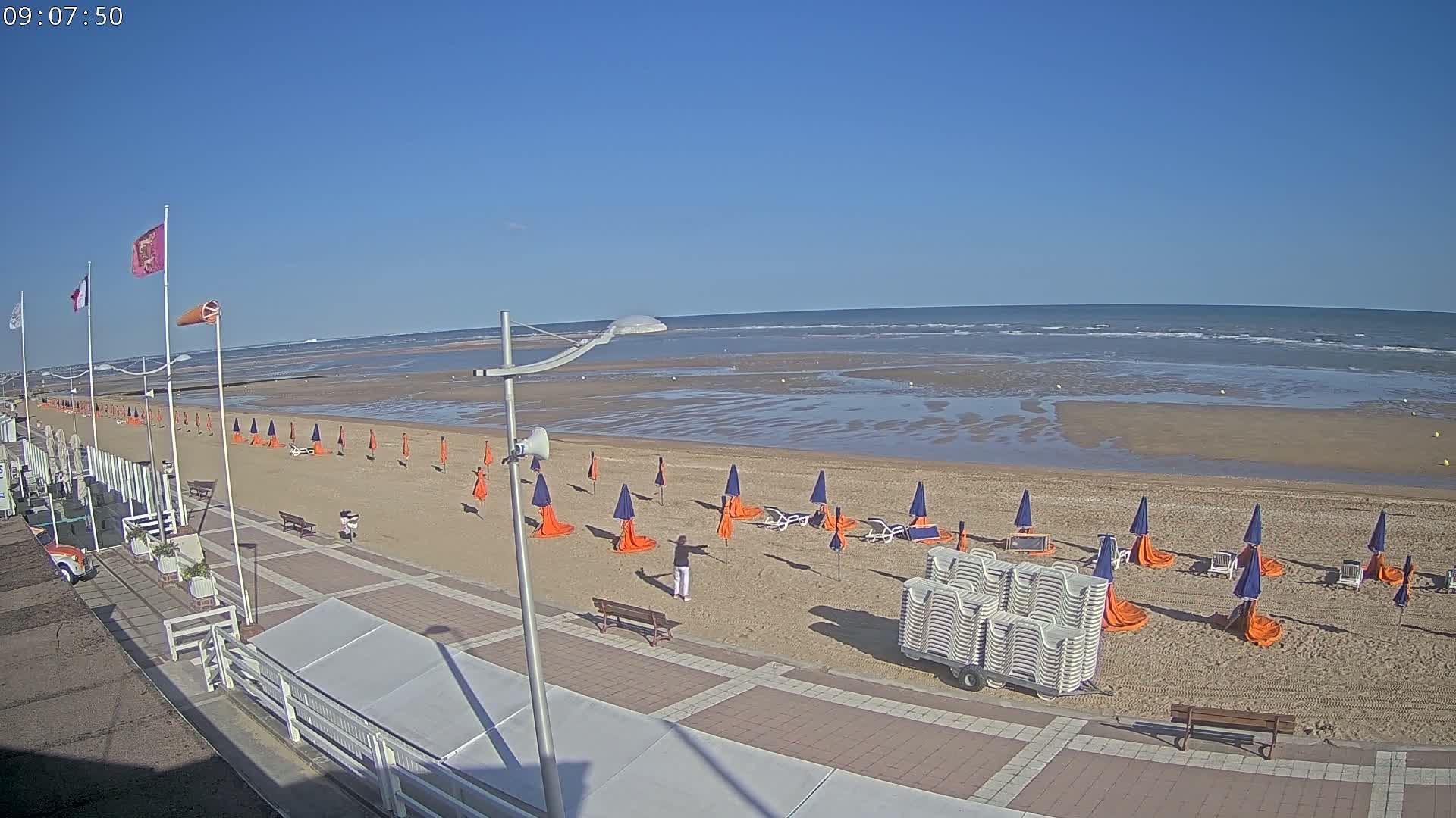 A mostly sunny beach scene shows rows of orange and blue beach umbrellas and chairs set up on the sand with a boardwalk and buildings in the foreground.