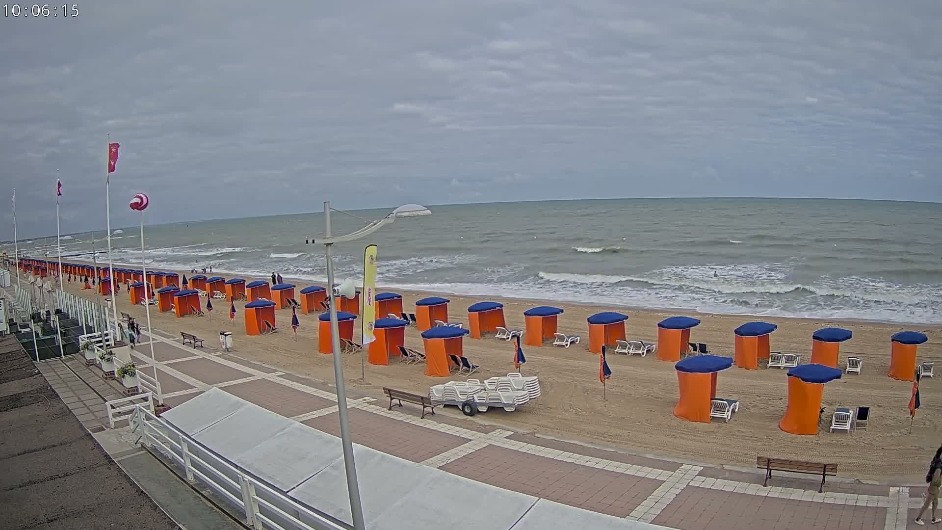A sandy beach with rows of orange and blue beach cabanas, a boardwalk, and a somewhat choppy grey sea under a cloudy sky.