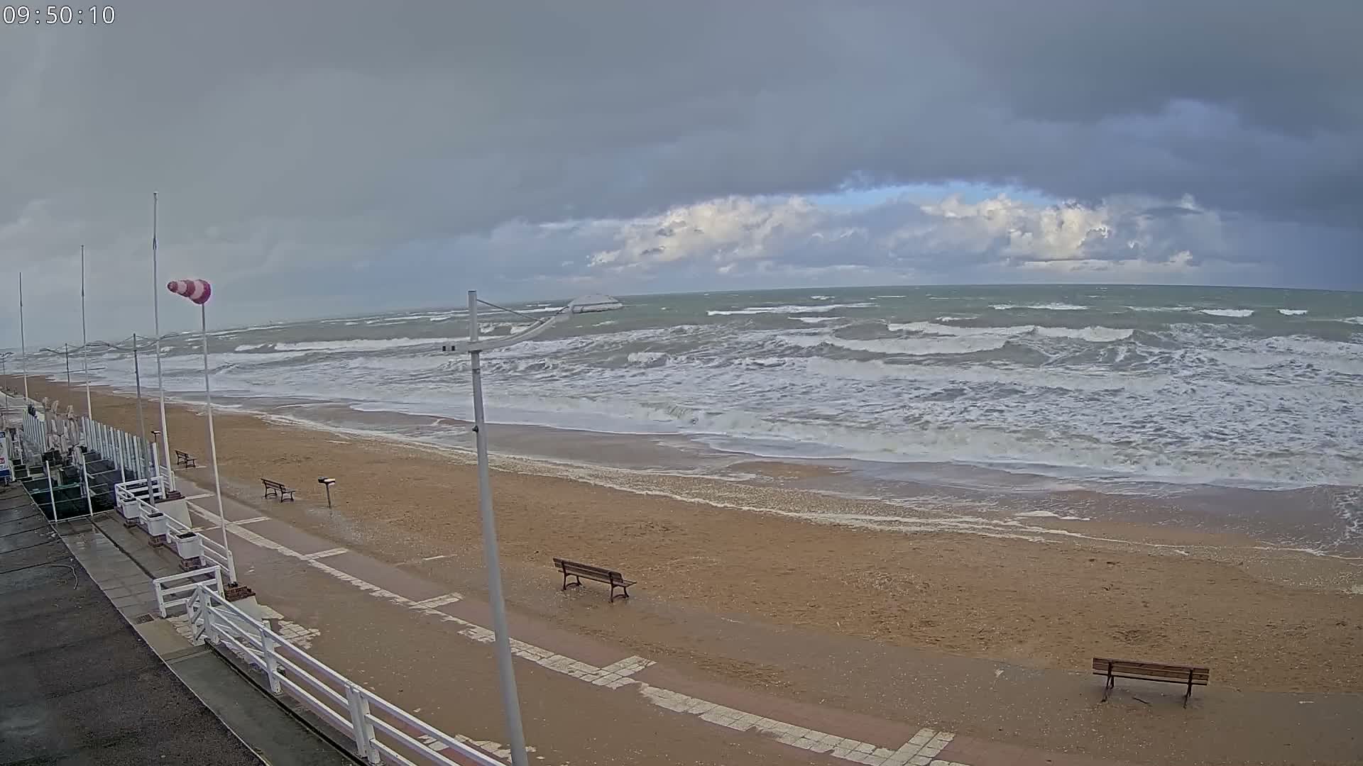 A deserted beach with strong waves crashing on the sandy shore and a windsock on the promenade is pictured under a heavily overcast, blustery sky.