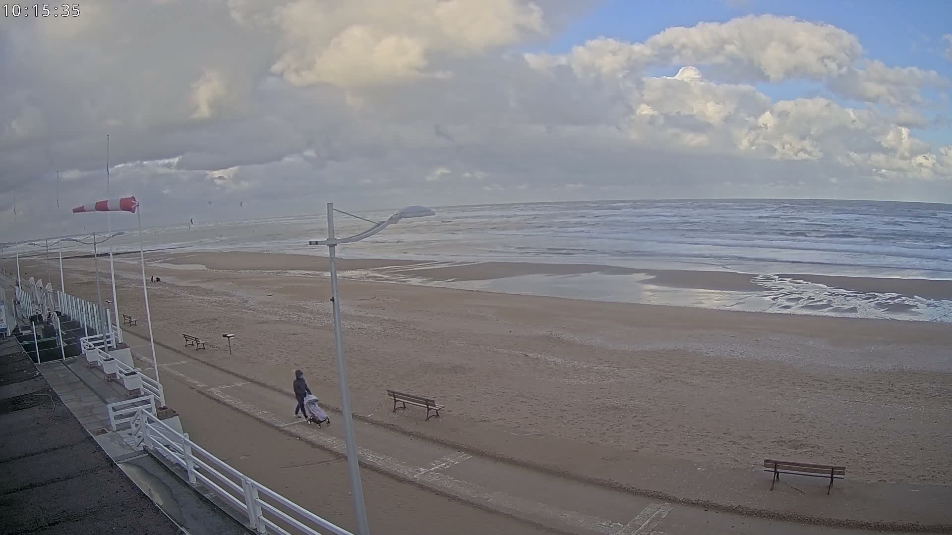 A windy, partly cloudy day at the beach features a person pushing a stroller along a sandy path beside the choppy ocean, with kite surfers visible in the distance.