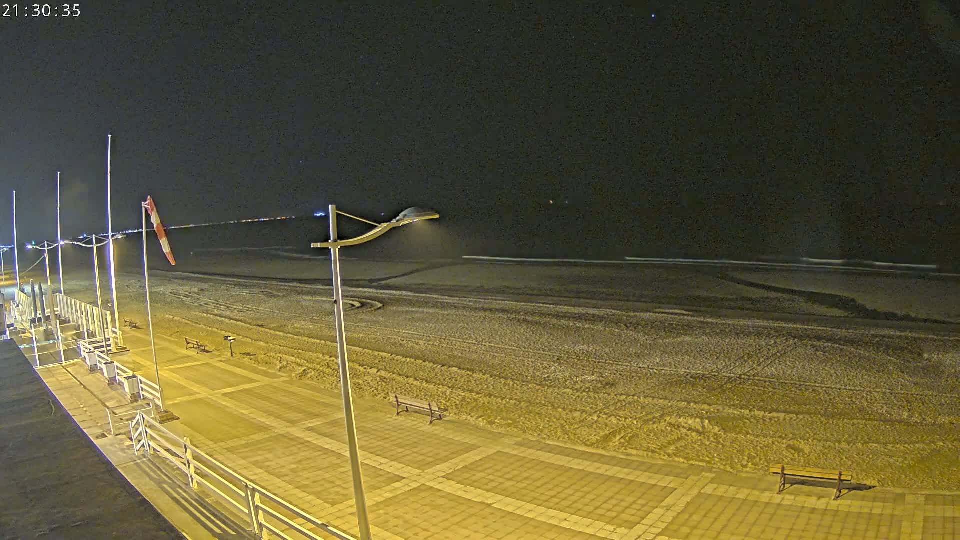 A windy, partly cloudy day at the beach features a person pushing a stroller along a sandy path beside the choppy ocean, with kite surfers visible in the distance.