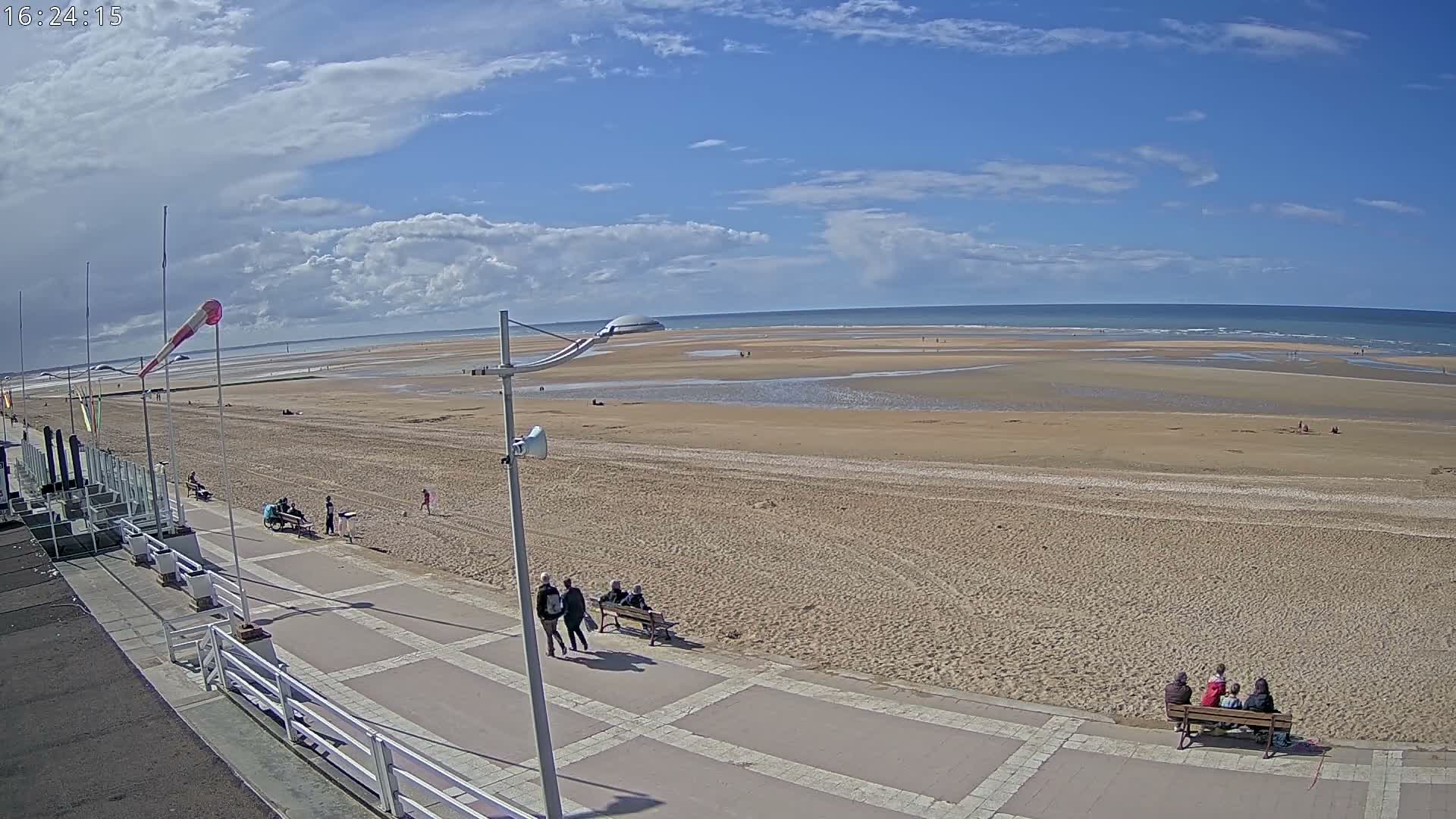 A windy, partly cloudy day at the beach features a person pushing a stroller along a sandy path beside the choppy ocean, with kite surfers visible in the distance.