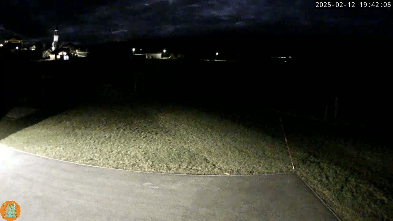 A nighttime view of a paved area next to a grassy hill, with a dimly lit town visible in the background under a partly cloudy sky.