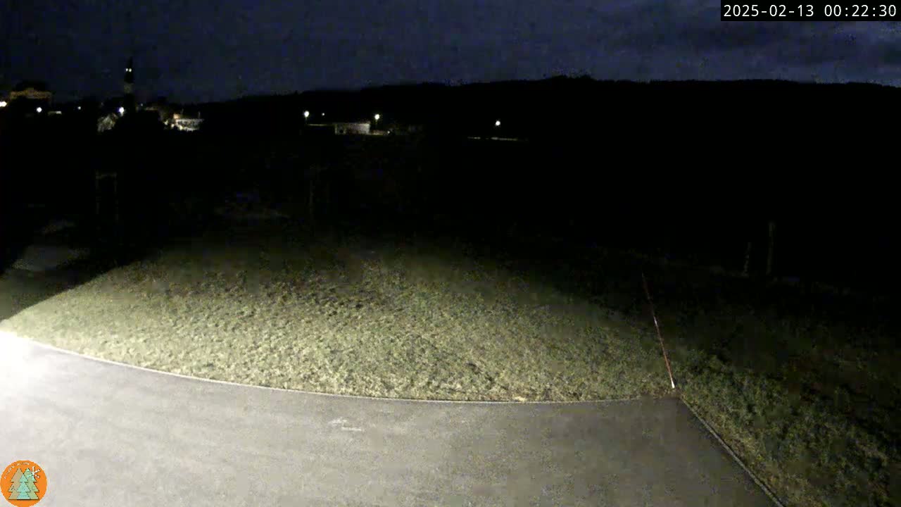 A nighttime view shows a dark, grassy area and a paved road curving into the foreground under a partly cloudy sky.
