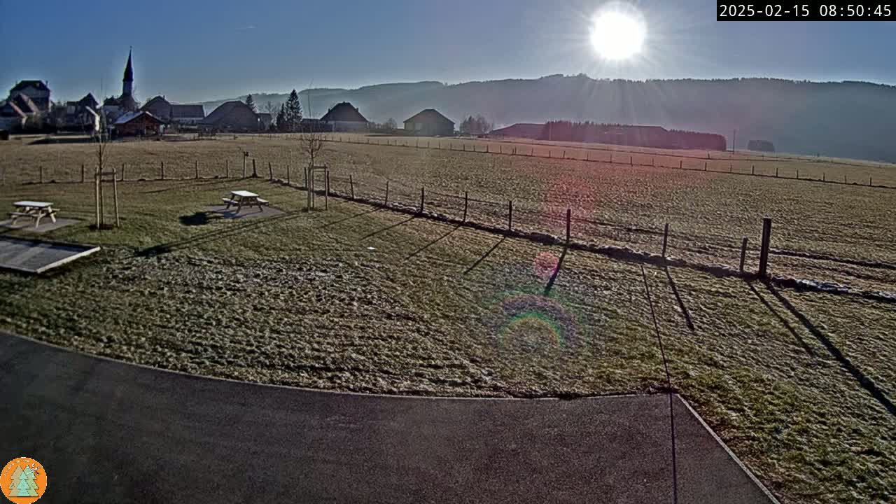 A frost-covered field with picnic tables and a small village in the background under a bright, sunny sky.