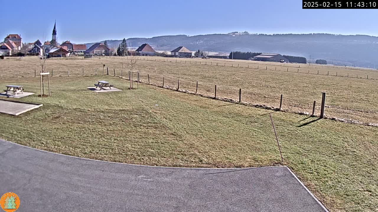 A paved area curves into a grassy field with picnic tables, a distant village, and mountains under a clear blue sky.