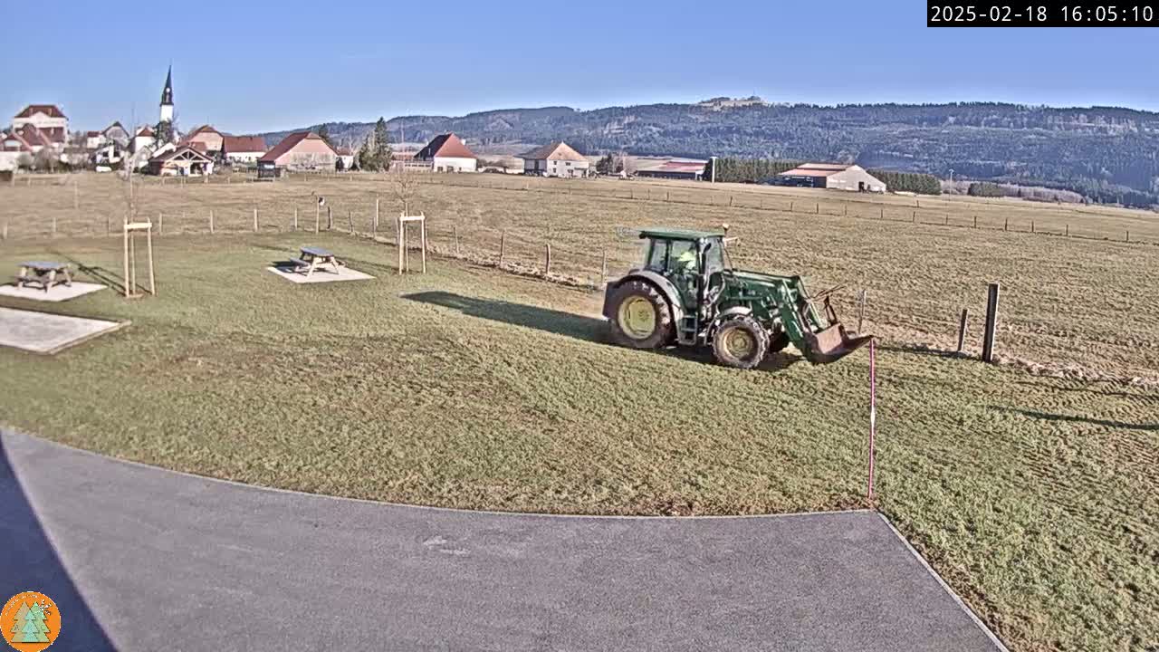 A green tractor with a bucket attachment is parked in a grassy field on a sunny day, near a paved area with picnic tables and a small village in the background.