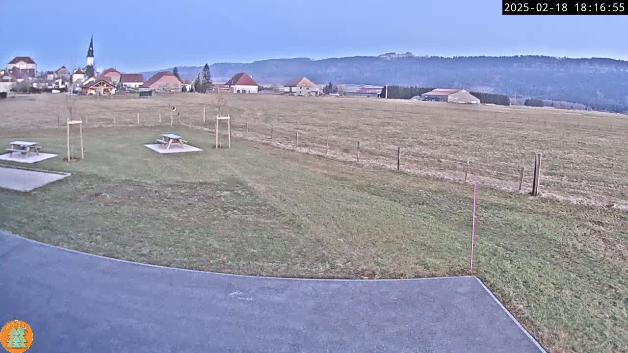 A paved area with two picnic tables overlooks a grassy field, beyond which sits a small village under a clear twilight sky.