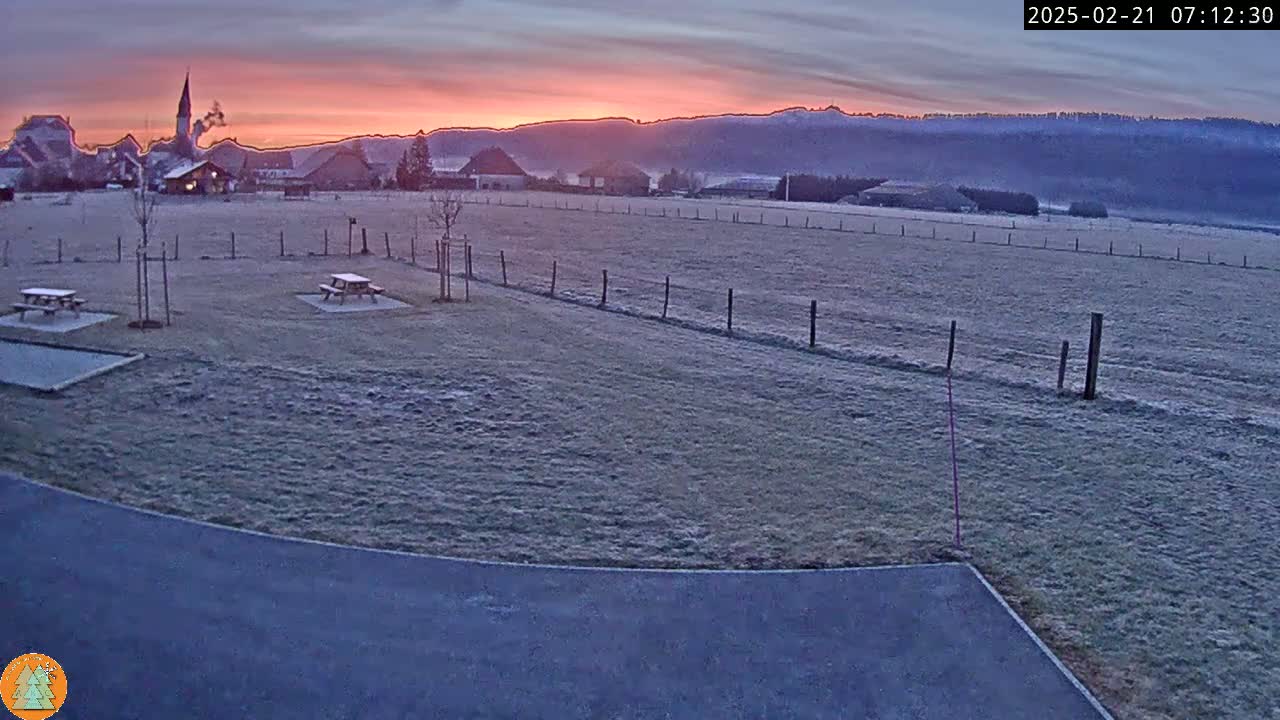 A frosty field with two picnic tables sits at sunrise before a small village and distant mountains.
