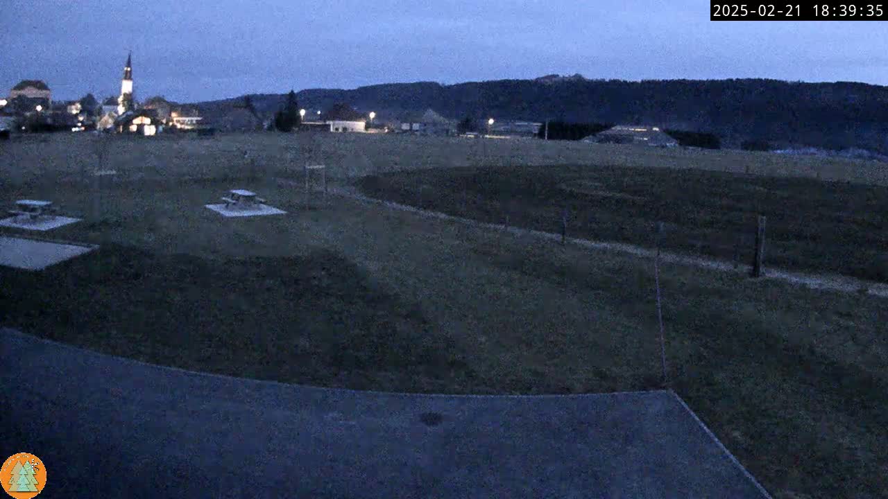 A dark, empty field with a few picnic tables and a small village in the distance under a twilight sky.
