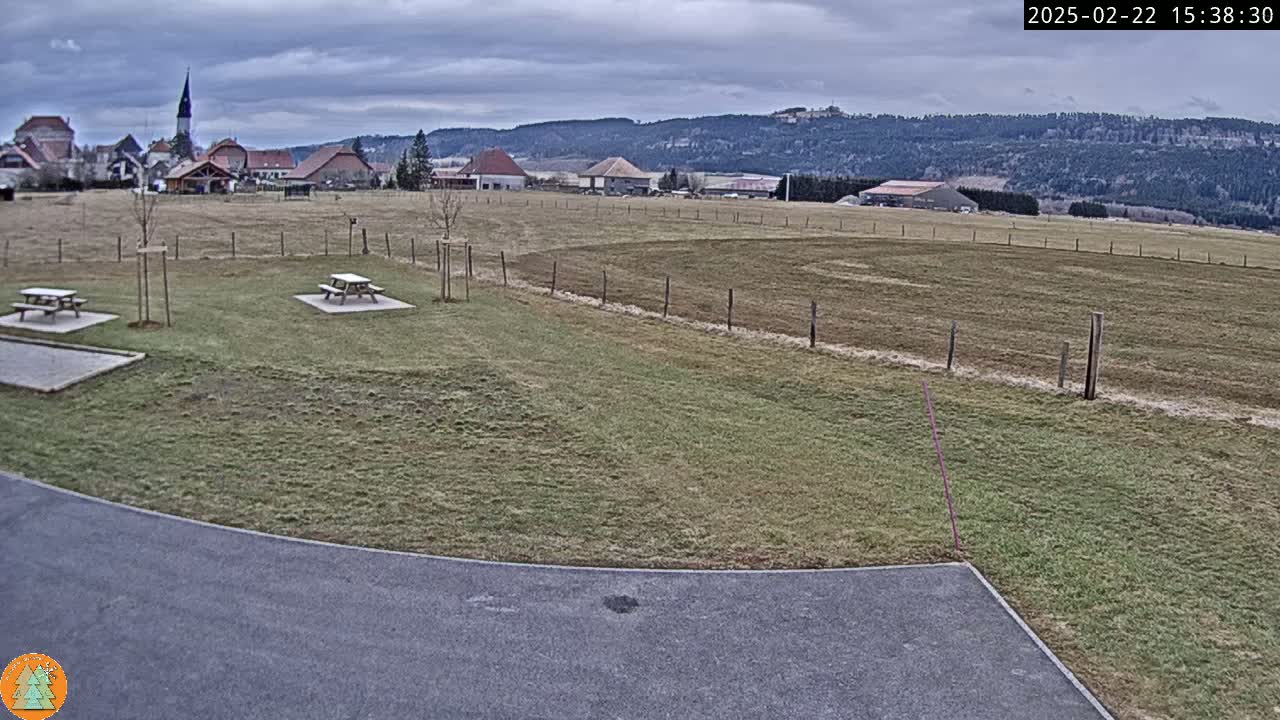 A mostly cloudy day reveals a grassy field with picnic tables, a paved area, and a village in the distance, nestled against a backdrop of hills and forests.