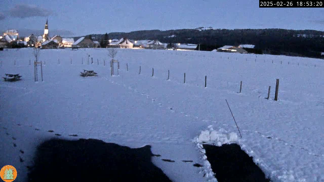 A snow-covered field with footprints, a few picnic tables, and a small village in the background under a twilight sky.
