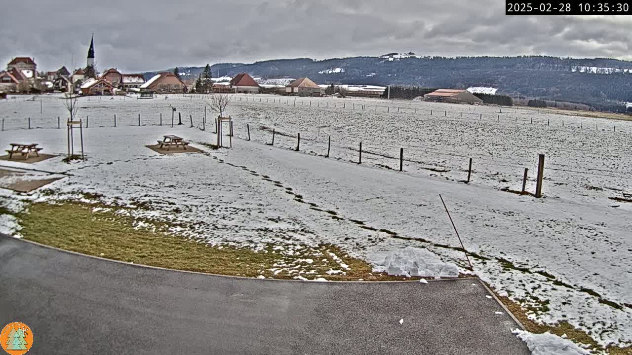 A snow-covered field with picnic tables and a small village in the background under a cloudy sky.