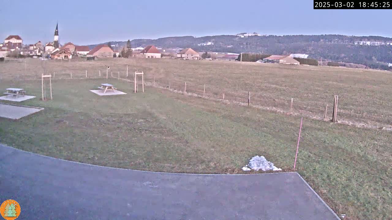 A grassy field with two picnic tables and a small village in the background under a clear twilight sky.