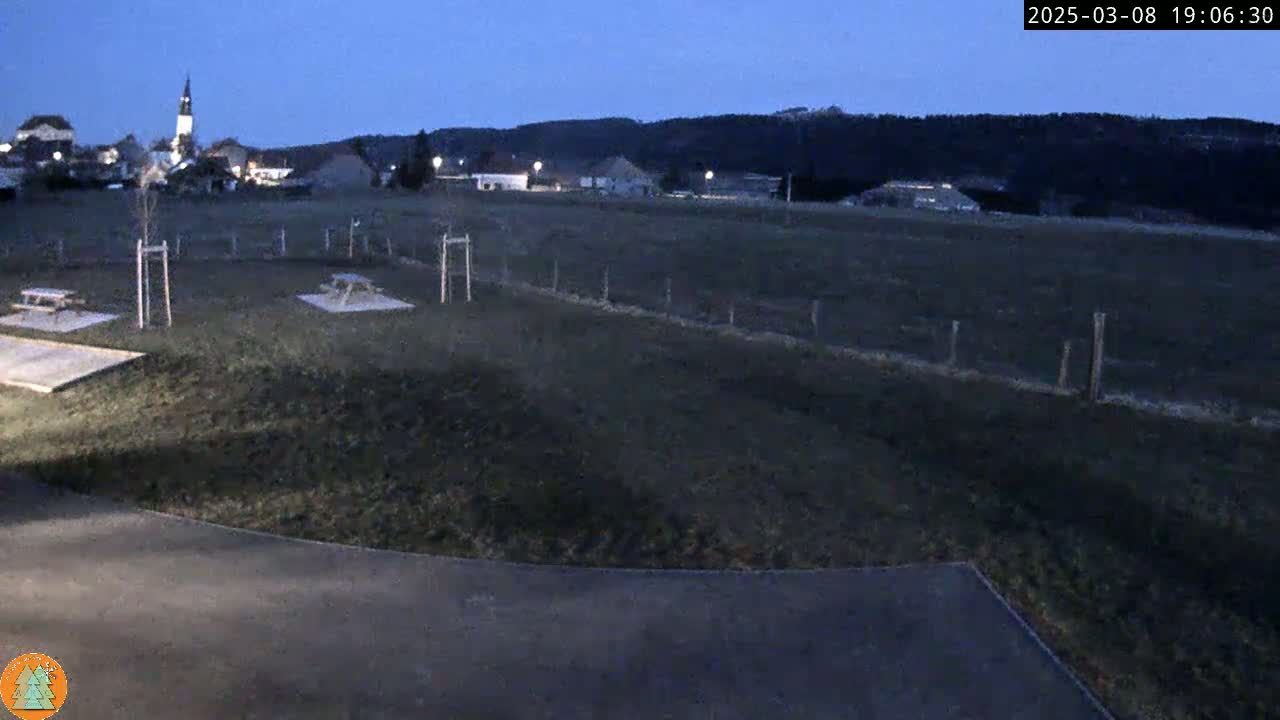 A dusk-lit, grassy field with picnic tables and a small village in the background under a clear, dark blue sky.