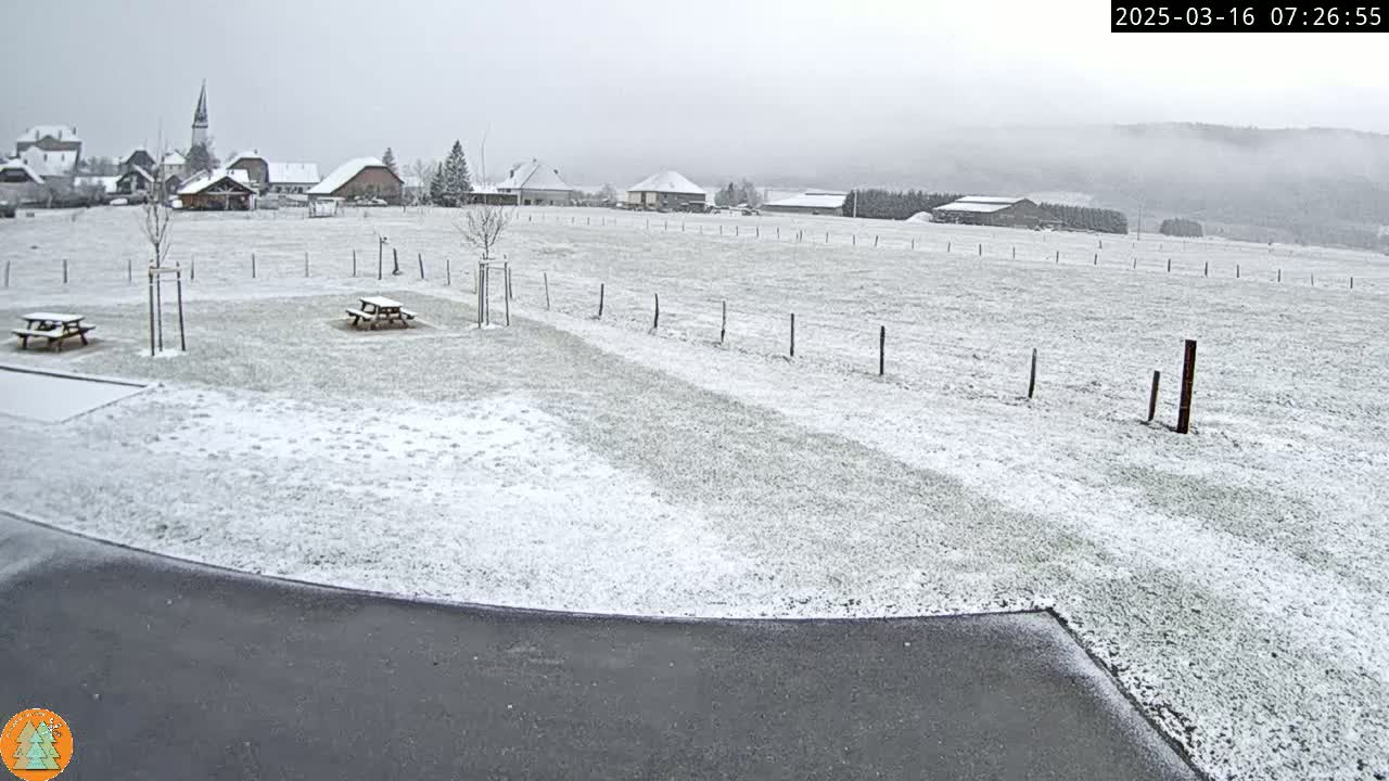 A snow-covered field with picnic tables and a small village in the background under an overcast sky.