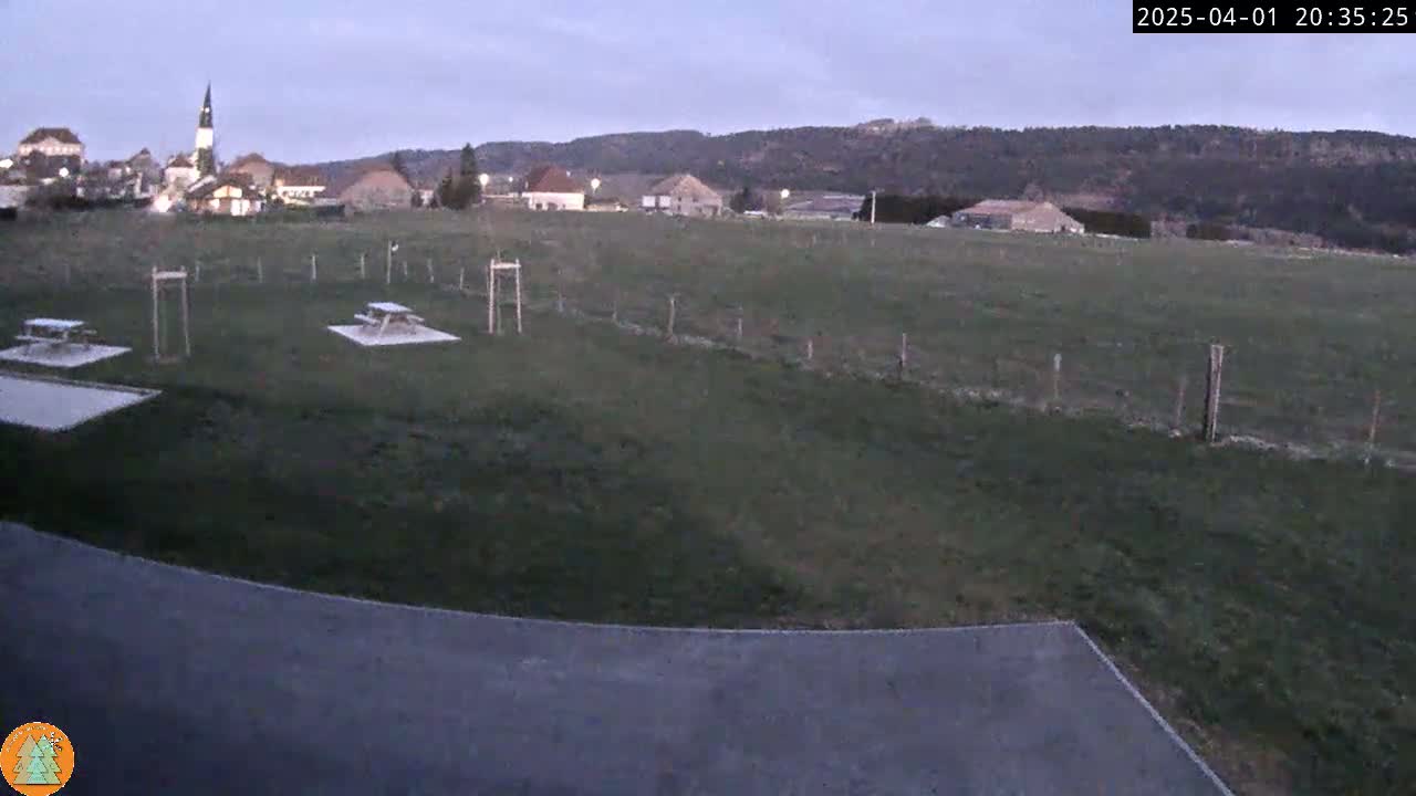 A grassy field with two picnic tables, a paved area, and a small village with a church steeple visible in the distance under a twilight sky.