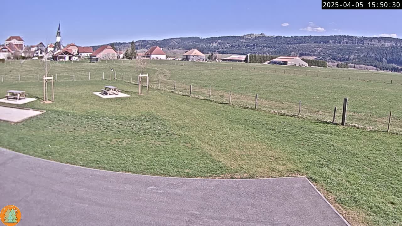 A grassy field with picnic tables sits in front of a small village nestled in a valley under a clear blue sky.