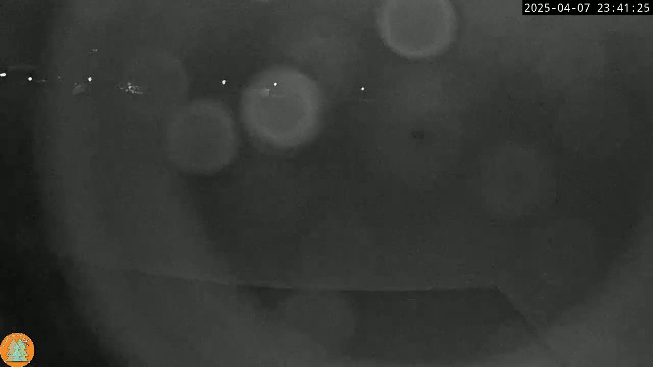 A blurry nighttime image shows out-of-focus lights in the distance, possibly from a road, with some light rain or condensation on a lens.
