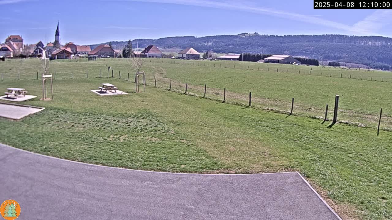 A grassy field with two picnic tables and a paved area in the foreground, under a clear blue sky, overlooks a small village nestled in a valley at the foot of a forested hill.