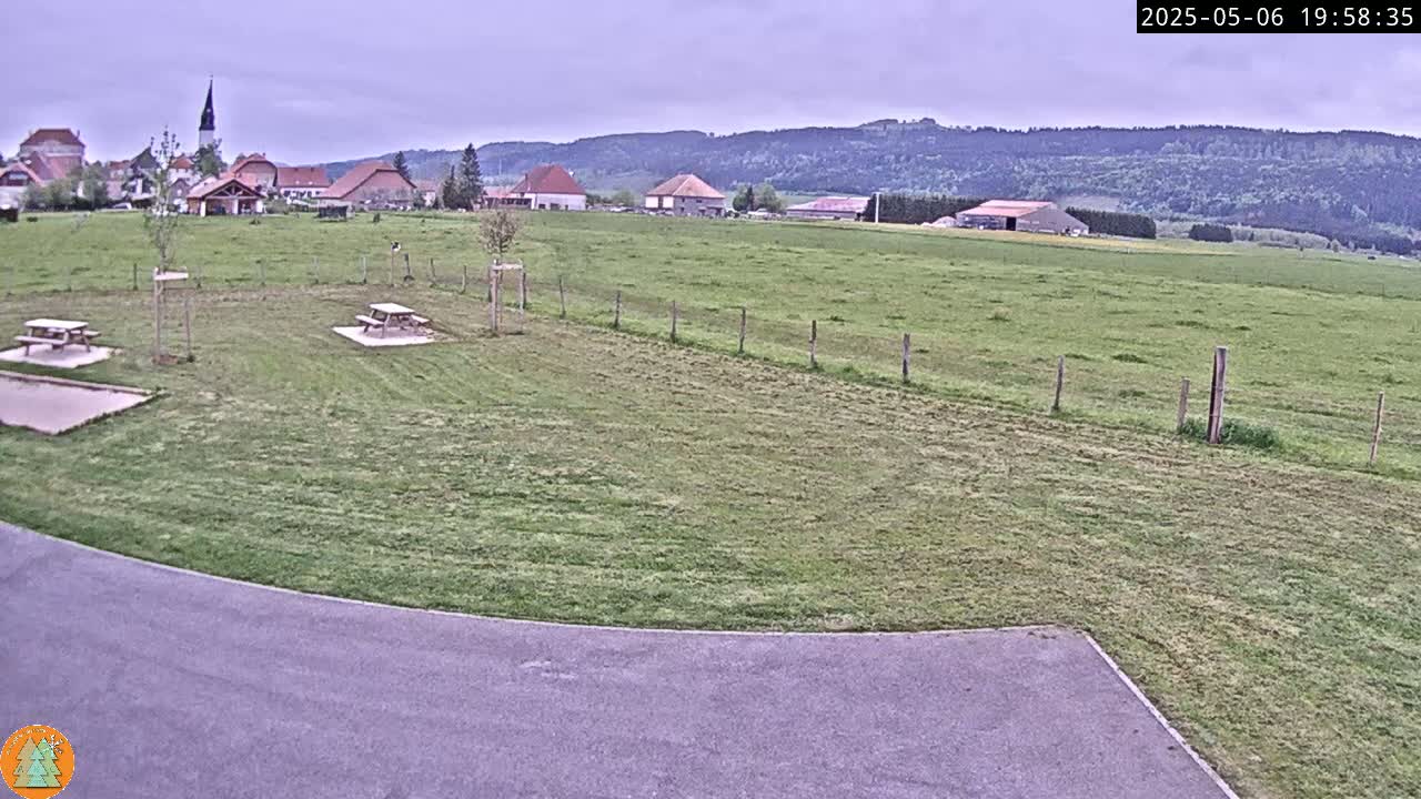 A grassy field with picnic tables sits in front of a small village under an overcast sky, with rolling hills visible in the background.