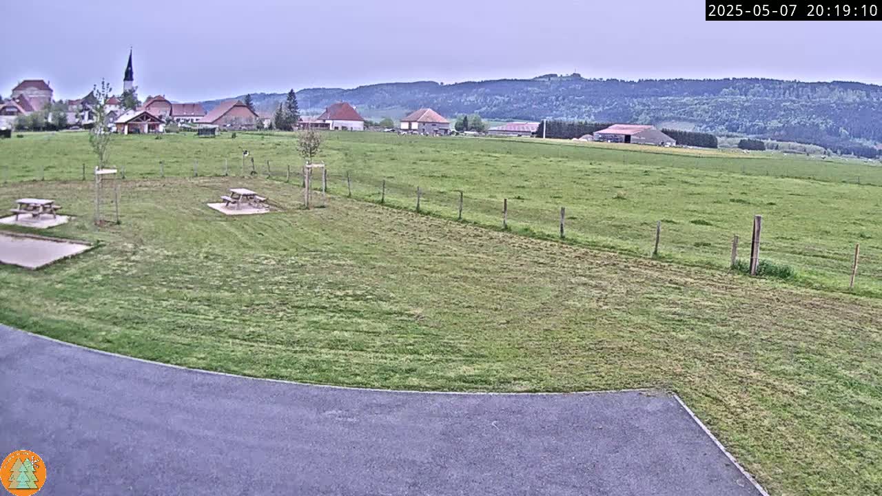 A grassy field with two picnic tables is in the foreground, overlooking a small village nestled against a range of hills under an overcast sky.