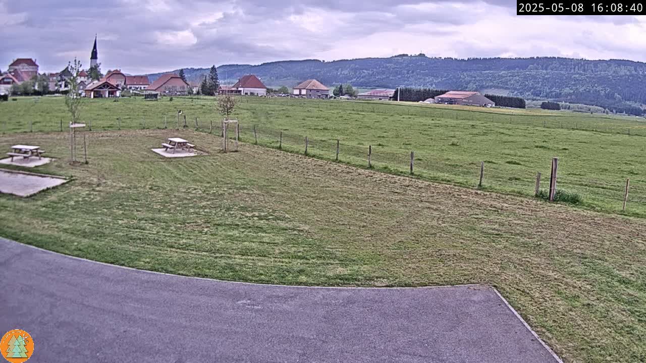 Under a partly cloudy sky, a grassy field with two picnic tables is situated in front of a small village nestled against a backdrop of rolling hills and forest.