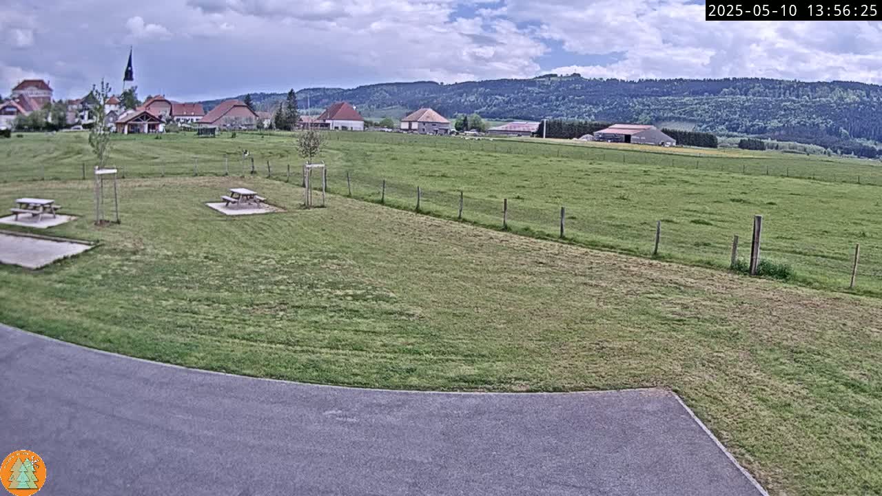 A partly cloudy day reveals a grassy field with two picnic tables, a paved area, and a small village nestled against a backdrop of rolling hills and forest.