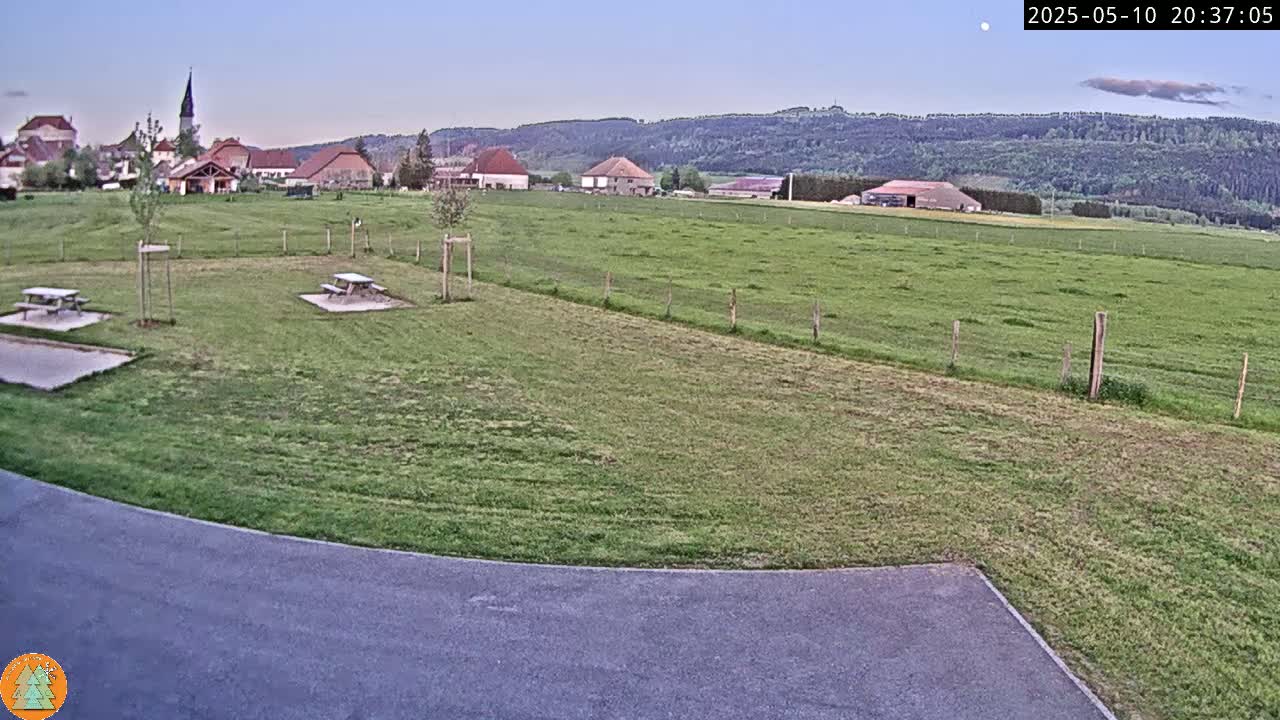 A grassy field with picnic tables sits in front of a small village nestled against a low mountain range under a twilight sky.