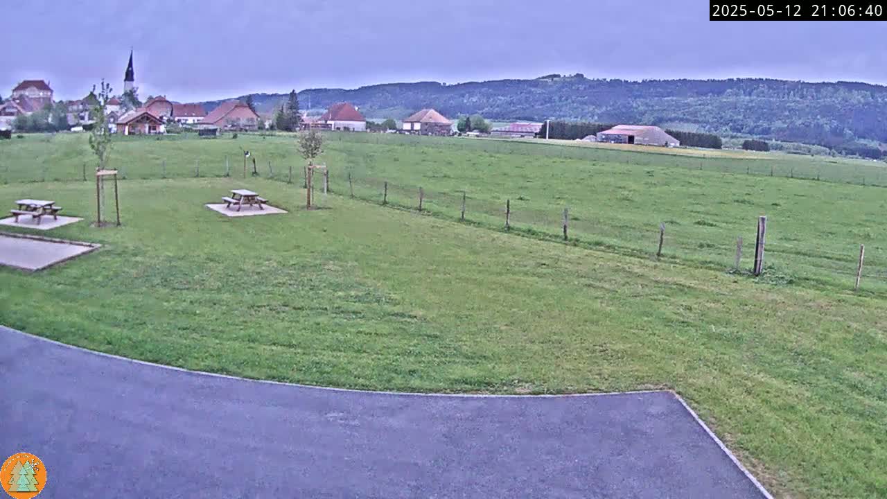 Two picnic tables sit on a grassy area overlooking a village nestled amongst rolling green hills under an overcast sky.