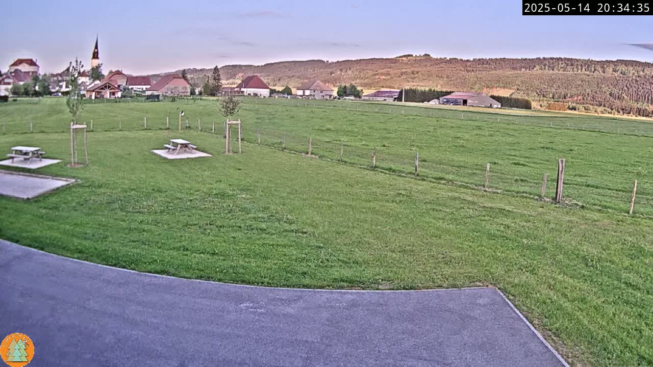 A grassy field with two picnic tables sits before a small village nestled at the base of a hillside under a twilight sky.