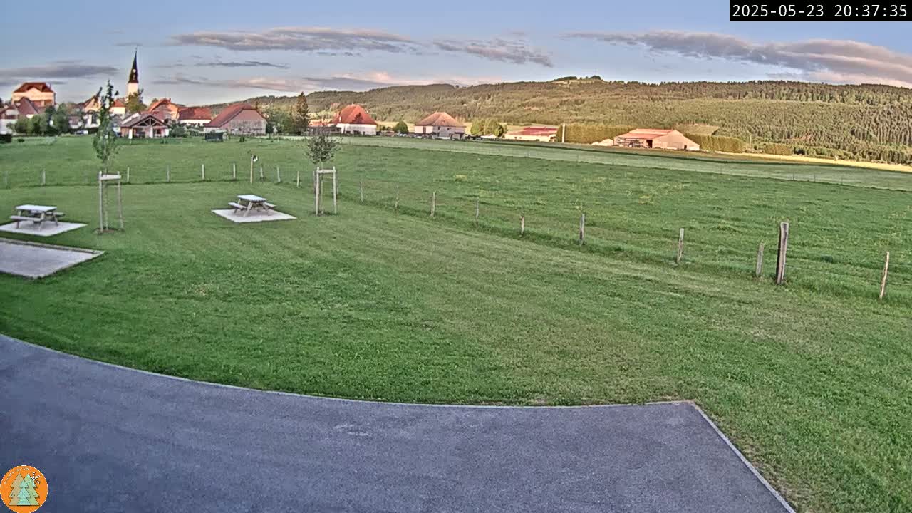 A grassy field with two picnic tables, a paved area, and a village with a church steeple in the background under a partly cloudy evening sky.
