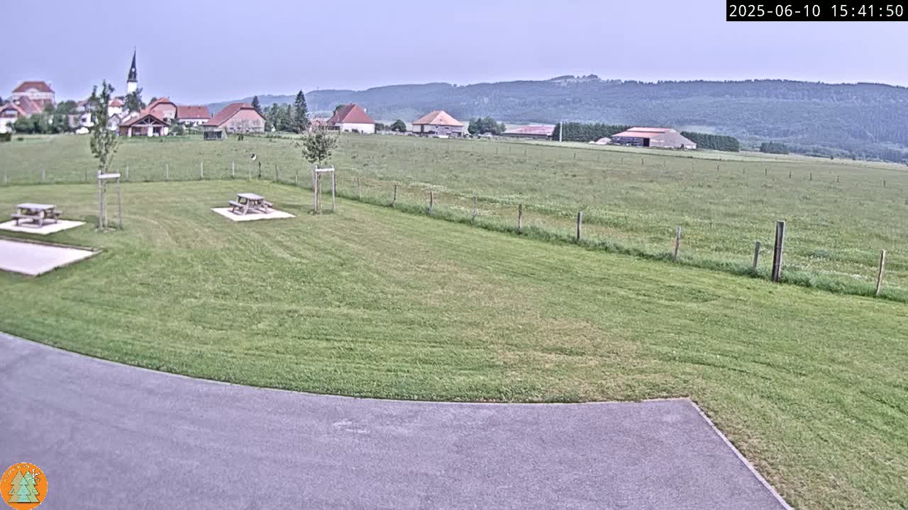 A grassy field with picnic tables sits in the foreground, overlooking a small village and rolling hills under a mostly cloudy sky.