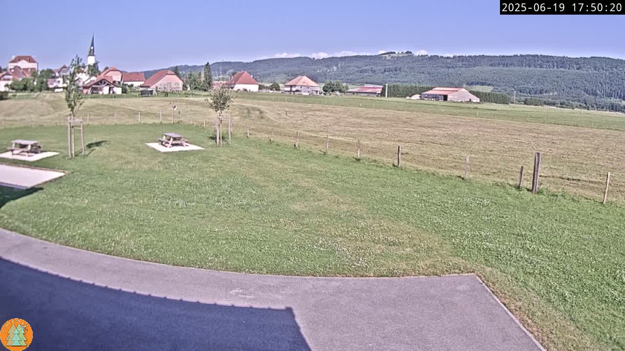 A grassy field with two picnic tables sits in the foreground, overlooking a small village nestled against a backdrop of forested hills under a clear blue sky.