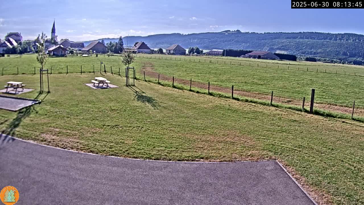 A grassy field with picnic tables sits in front of a small village nestled at the base of a line of forested hills under a clear, sunny sky.