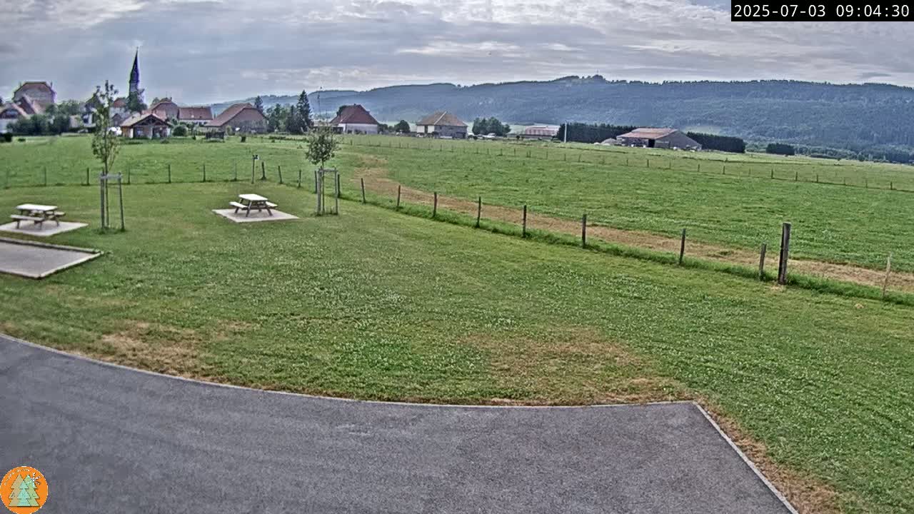 A grassy field with picnic tables and a paved area overlooks a small village nestled in a valley under a cloudy sky.