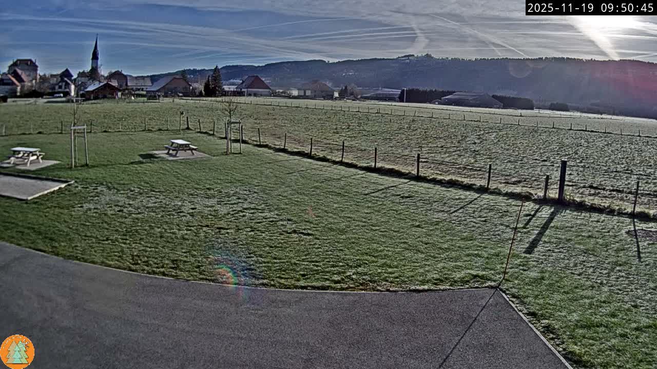 A wide view captures a frosty green field with two picnic tables and a gravel court in the foreground, leading to a distant village with a prominent church steeple and rolling hills under a bright, clear morning sky adorned with numerous contrails.