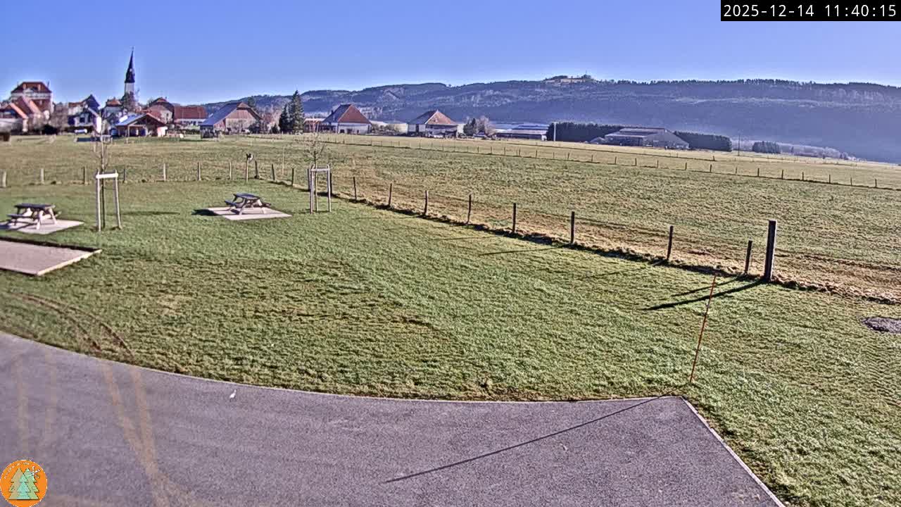 Under an overcast sky, a wide outdoor scene is blanketed in fresh snow, revealing a distant village with a prominent church steeple, fenced fields, and two snow-covered picnic tables.