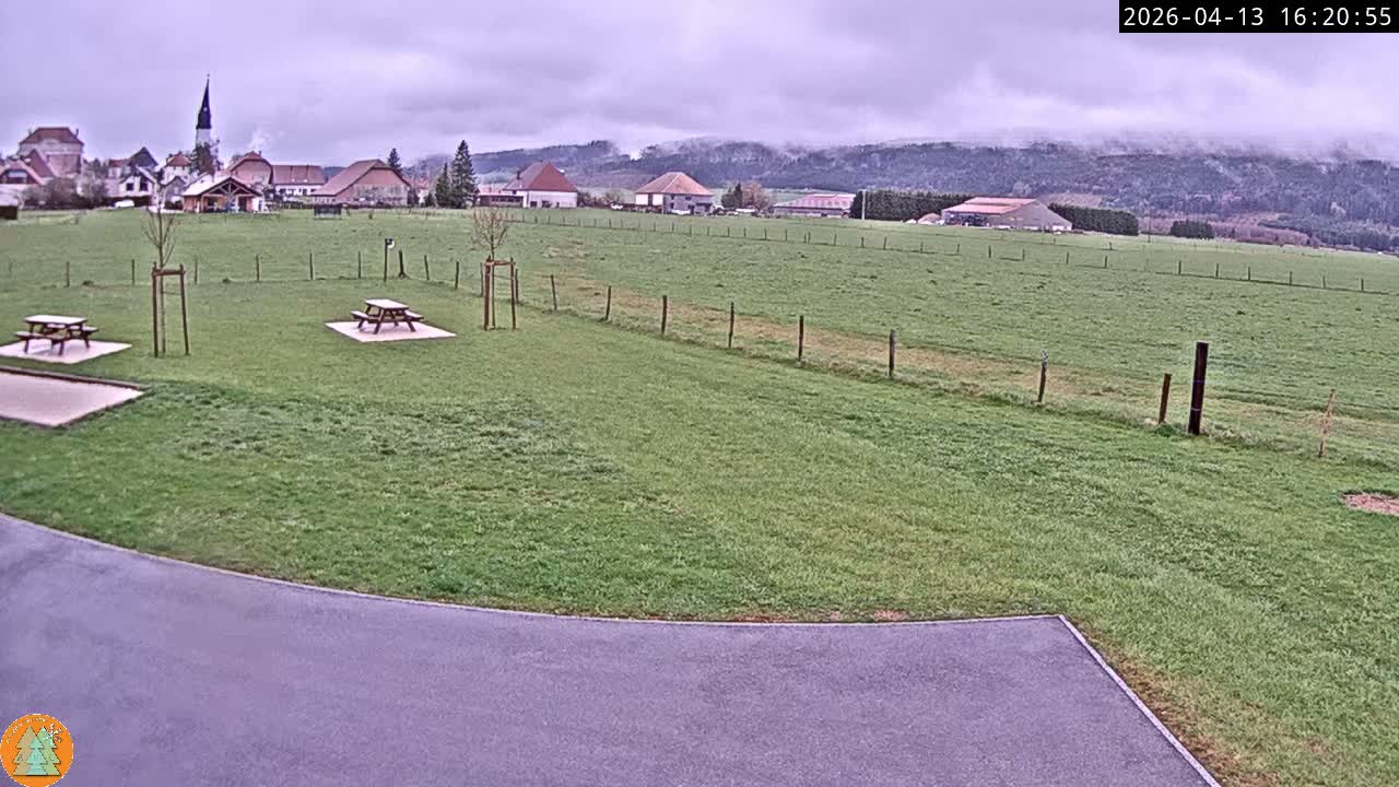 Under an overcast sky, a wide outdoor scene is blanketed in fresh snow, revealing a distant village with a prominent church steeple, fenced fields, and two snow-covered picnic tables.