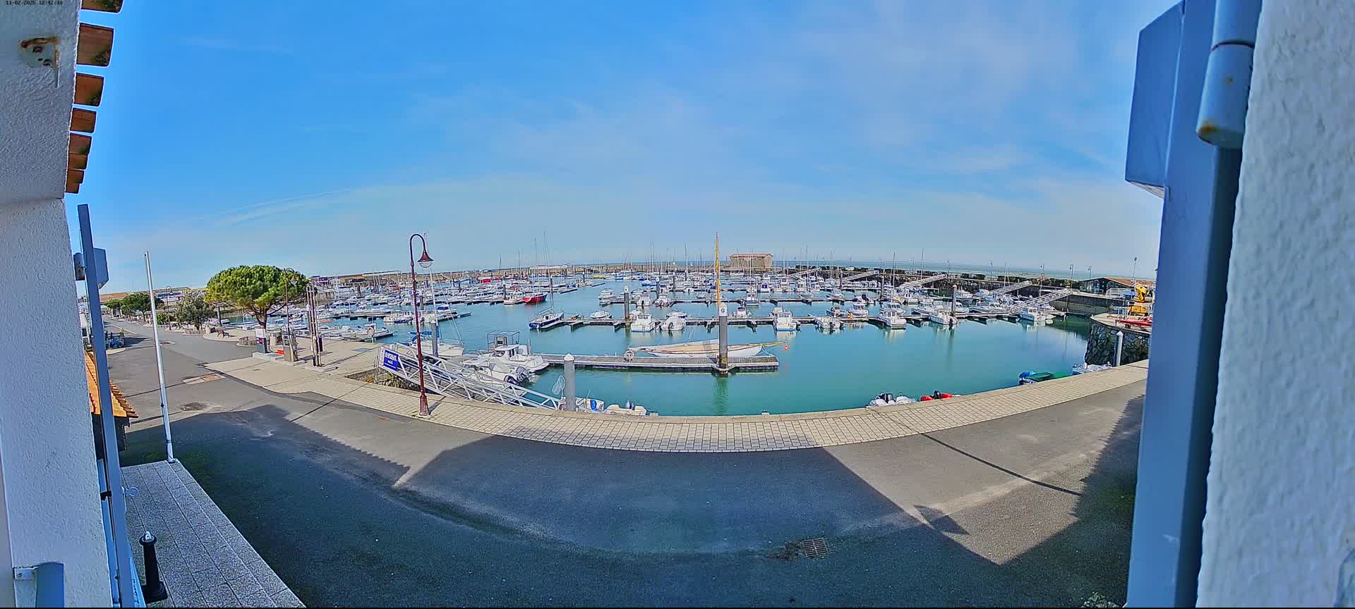 A sunny day reveals a calm harbor filled with numerous boats docked at various points, viewed from a slightly elevated perspective.