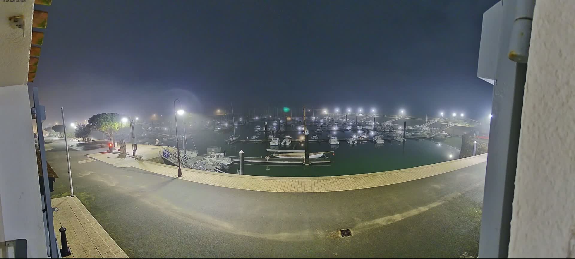 A fog-shrouded marina at night is illuminated by numerous lights, showing many boats docked in the calm water.