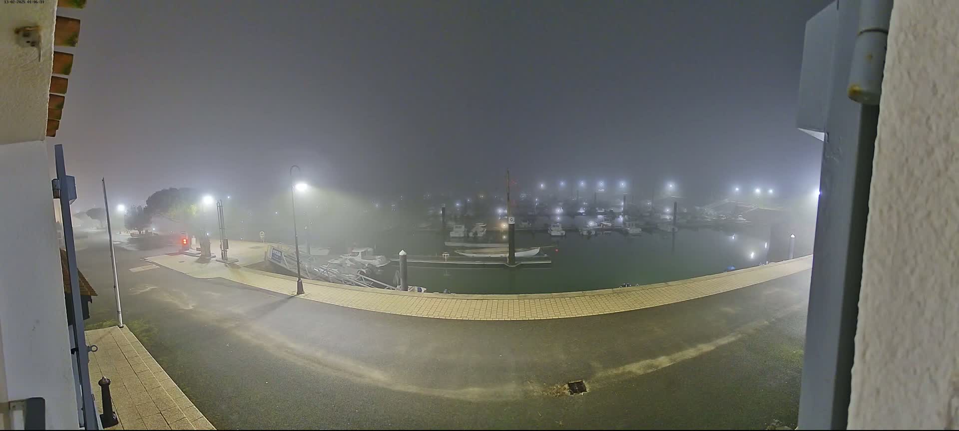 A foggy night scene shows a marina with numerous boats moored, illuminated by streetlights, viewed from a paved area.