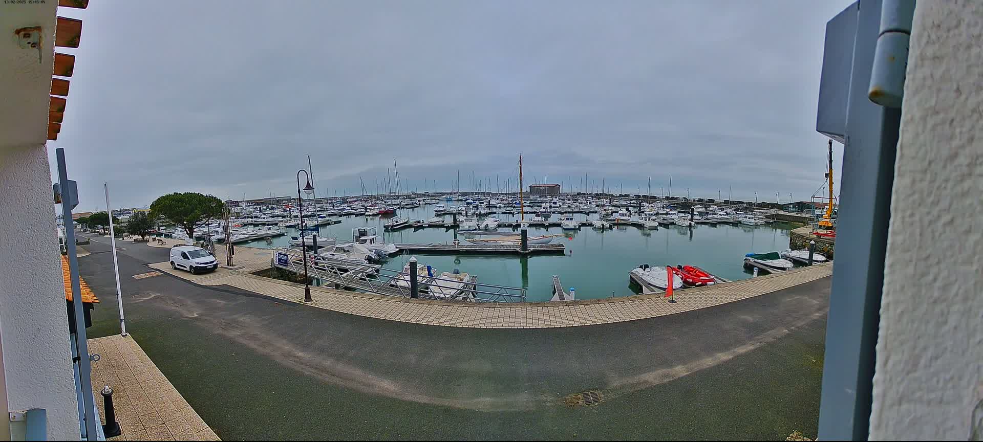 A marina filled with numerous boats is seen on an overcast day, with a single white van parked on the adjacent roadway.