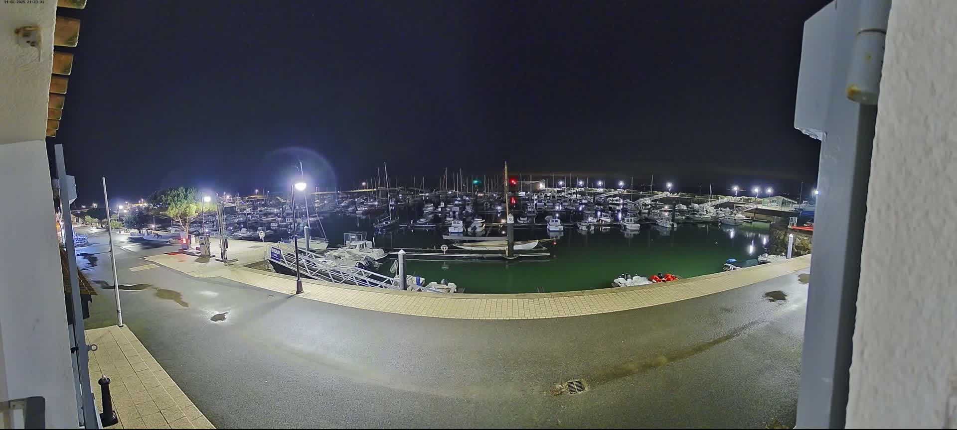 A nighttime view of a marina filled with boats, illuminated by lampposts, with a wet paved area in the foreground.
